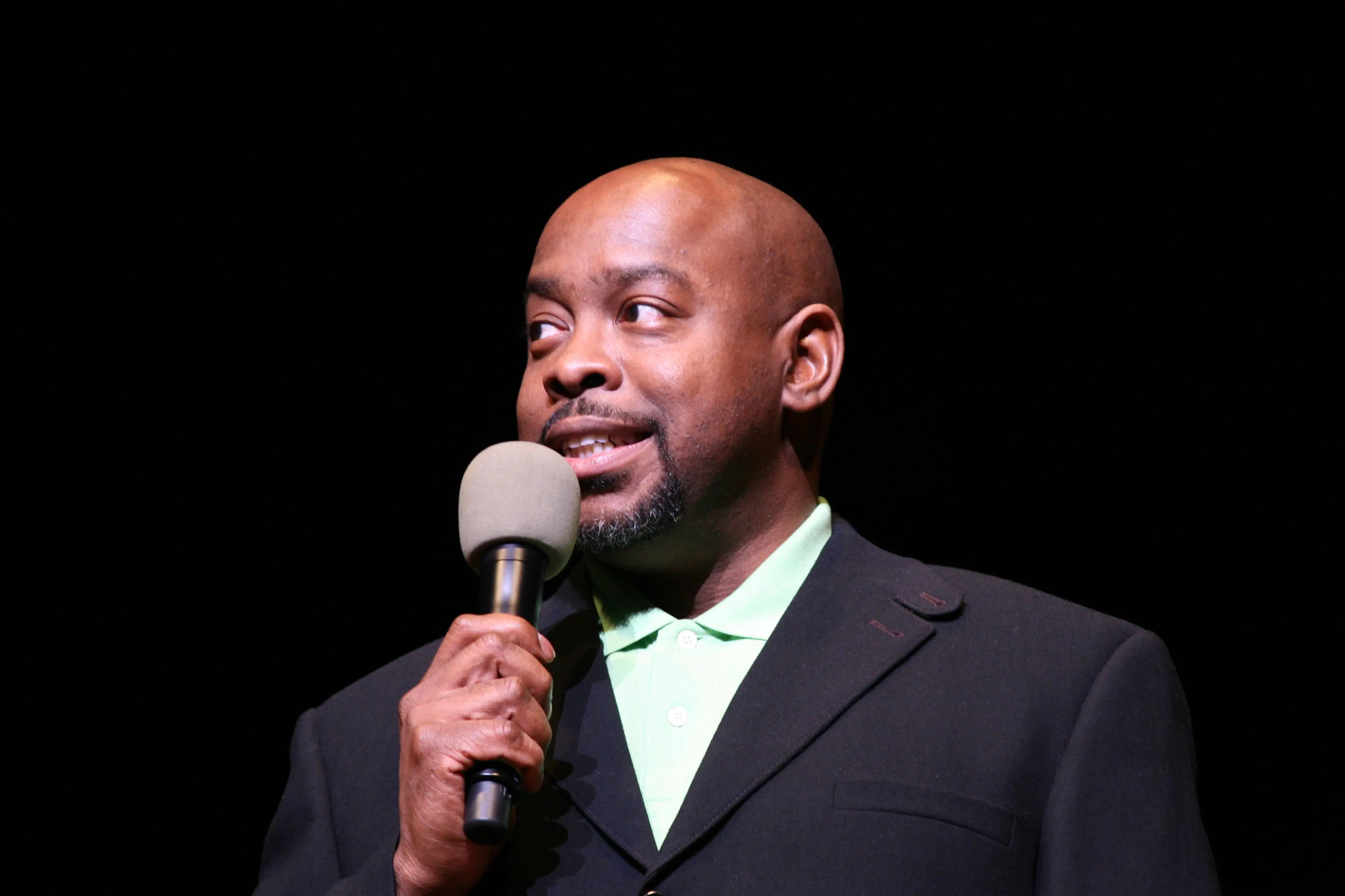 A performer in a dark suit speaking into a handheld microphone on an unlit stage