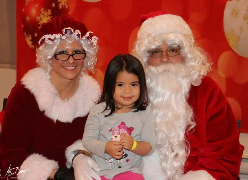 A young child on Santa's lap with Mrs Claus beside them at a holiday event