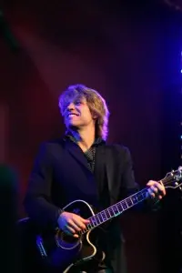 A guitarist smiling as he plays, lit blue and violet from the stage rig behind him
