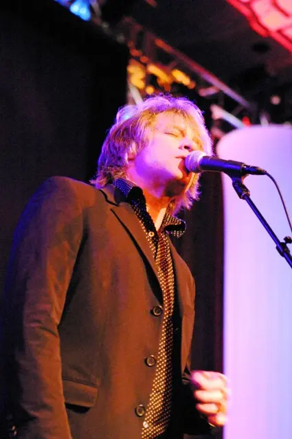 A singer at the microphone in a brown jacket, lit warm gold from above