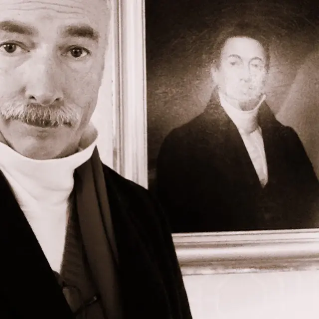 A man posing beside a framed antique oil portrait, matching the sitter's expression