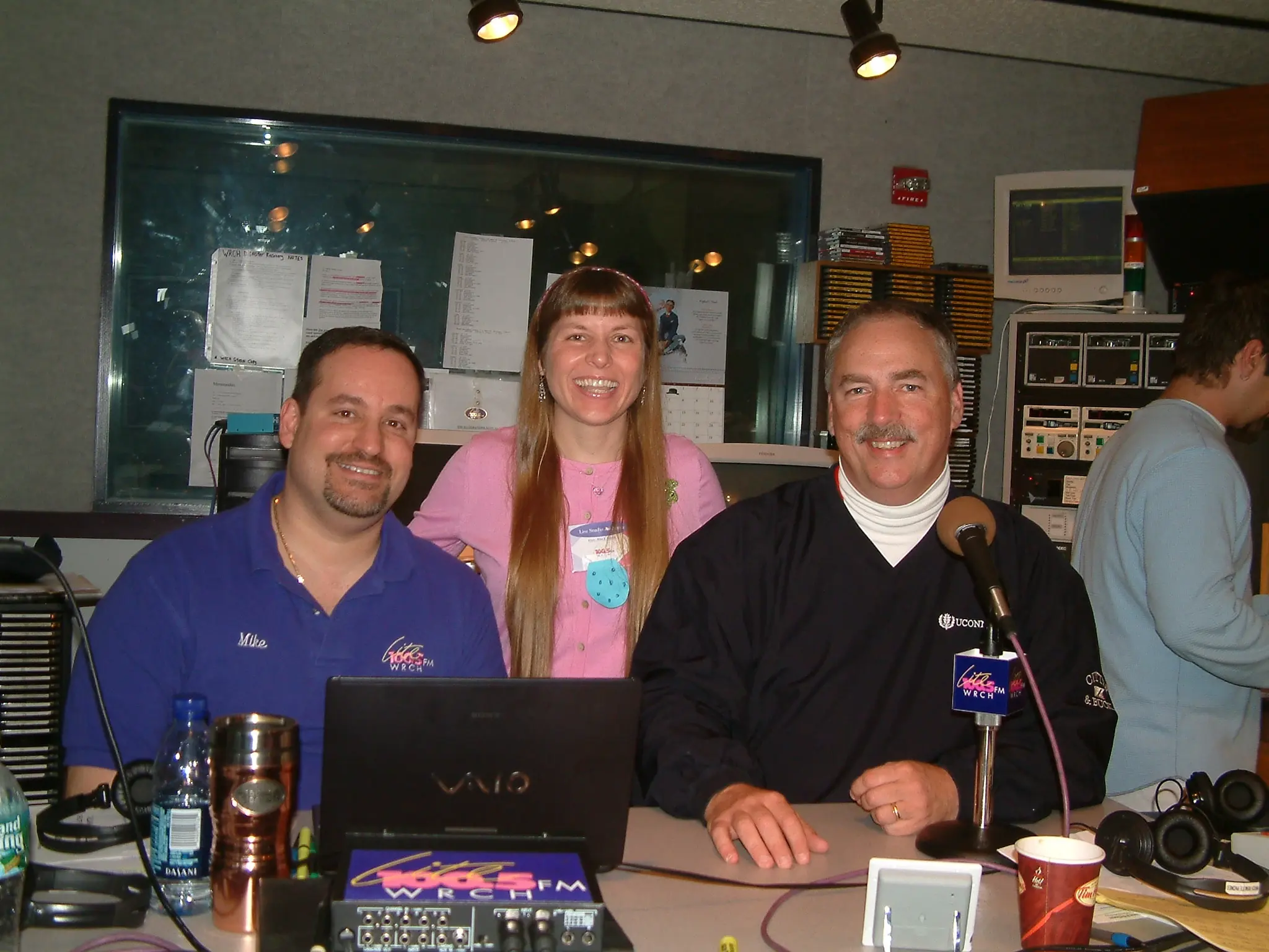 Three people behind a WRCH-FM remote broadcast desk, station microphone flag in front of them