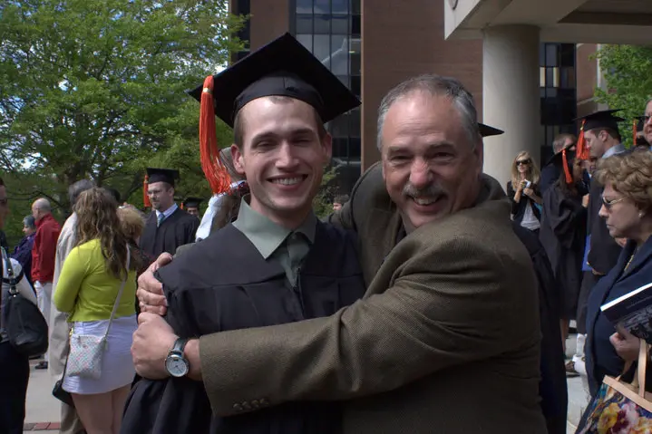 A graduate in cap and gown embraced by an older man outside a commencement ceremony