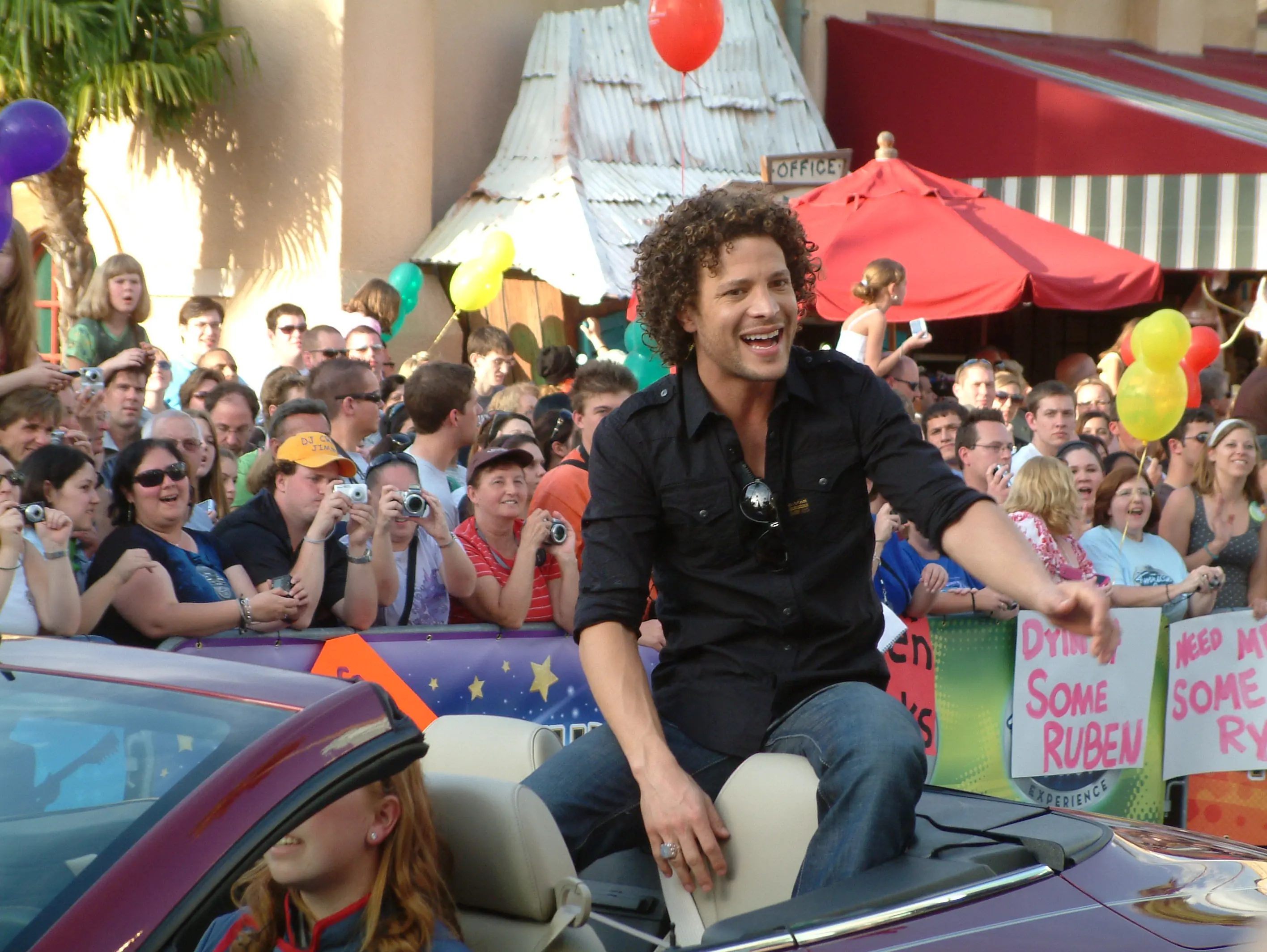 A performer perched on the back of a parade convertible, reaching toward a crowd of fans holding up cameras