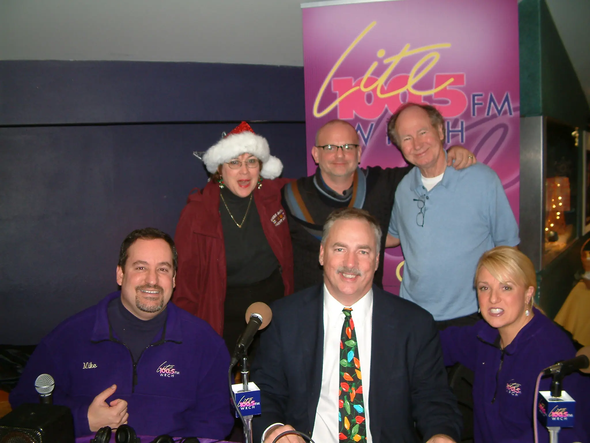 Station staff in purple Lite 100.5 jackets with guests, one wearing a Santa hat, in front of a Lite 100.5 FM WRCH banner