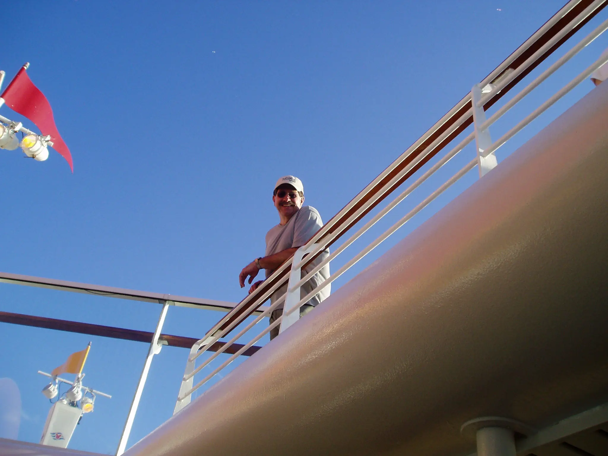 Looking up the side of a cruise ship at its rail and funnel against a clear blue sky, a passenger leaning on the rail