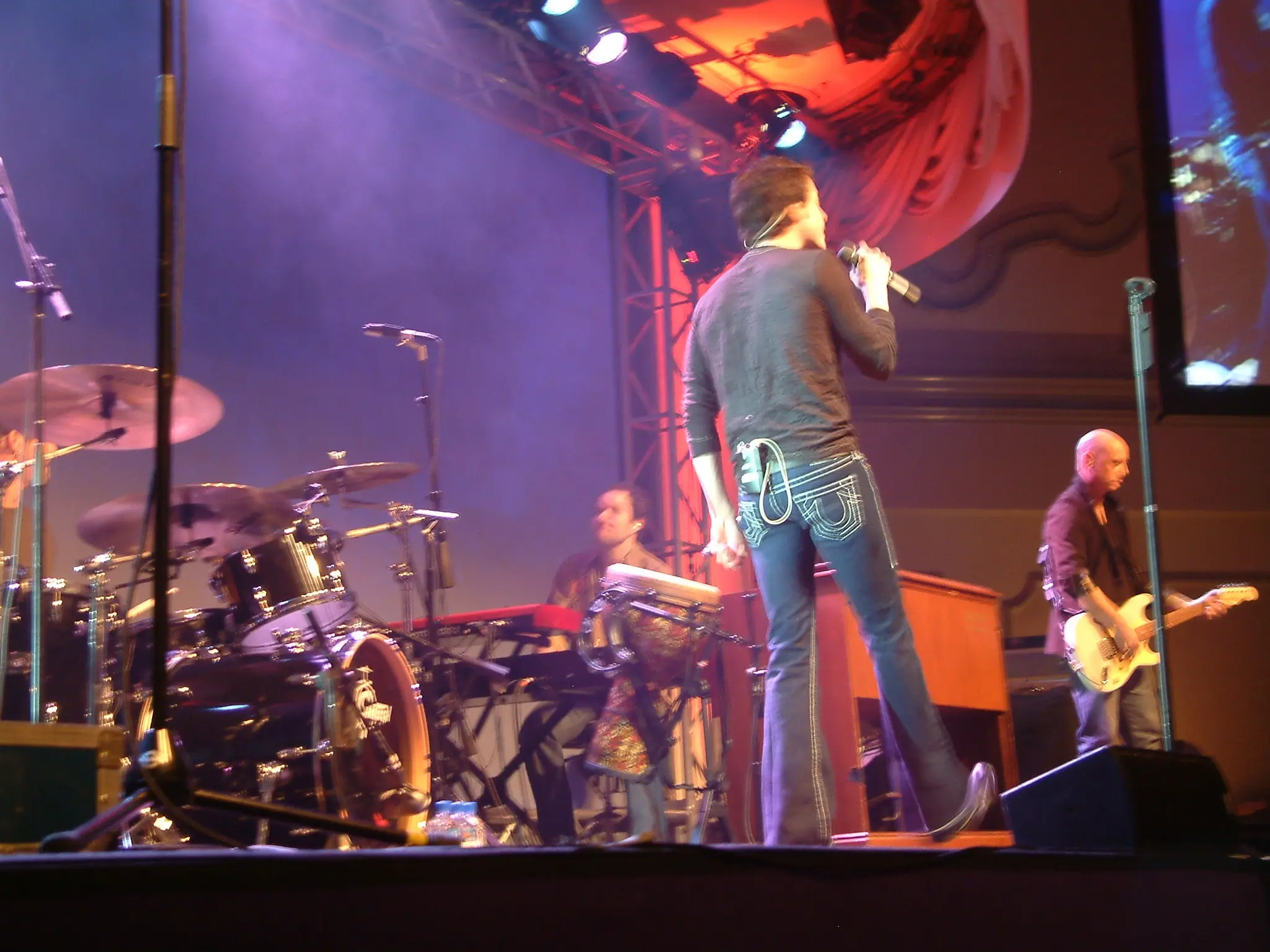A singer seen from behind on stage, facing the drum kit under magenta lighting