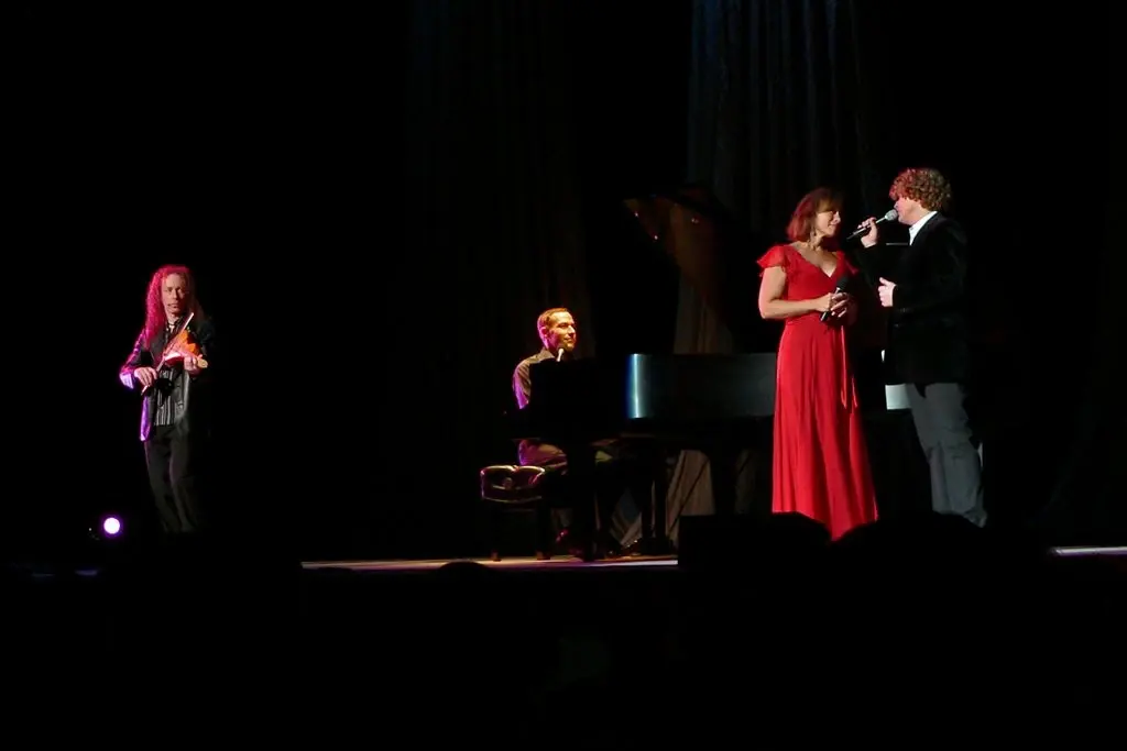 Two singers performing a duet beside a grand piano on a darkened stage, one in a long red gown