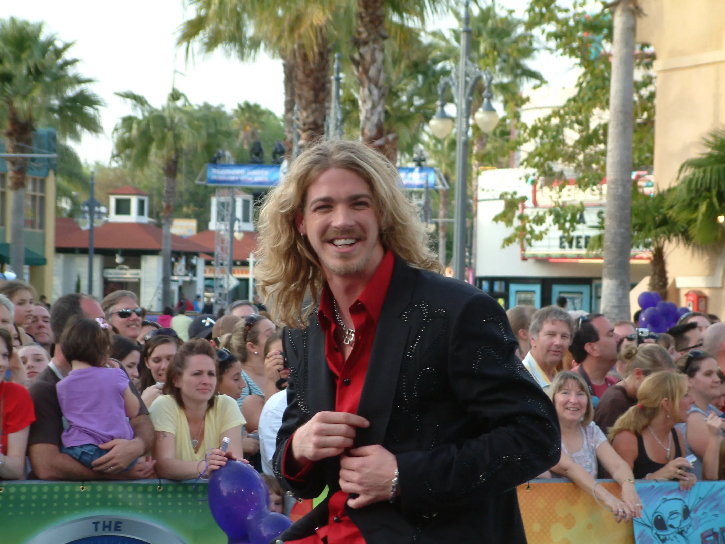 A long-haired performer in a black sequinned jacket and red shirt greeting fans lining a street