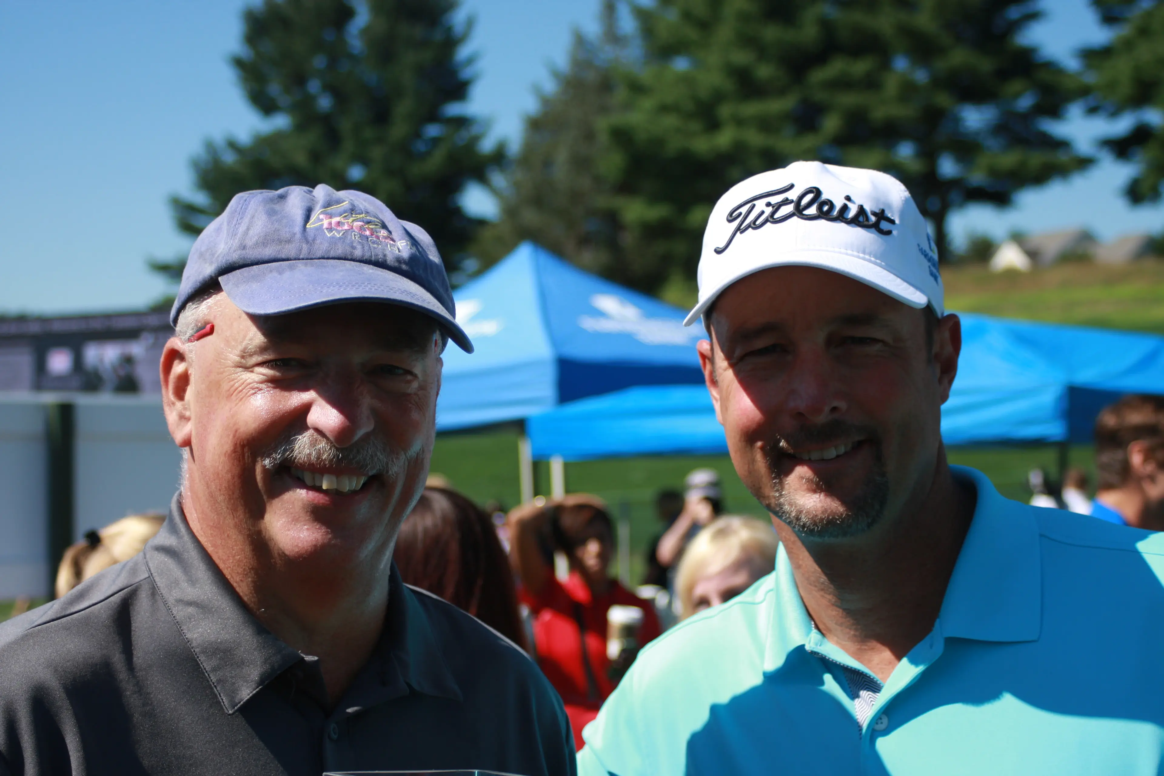 Two golfers in caps at an outdoor charity tournament, event tents behind them