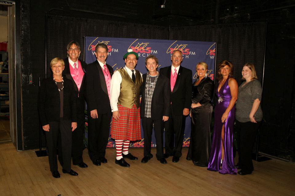 A group in formal wear, two of them in kilts, posed together in front of a Lite 100.5 FM WRCH backdrop