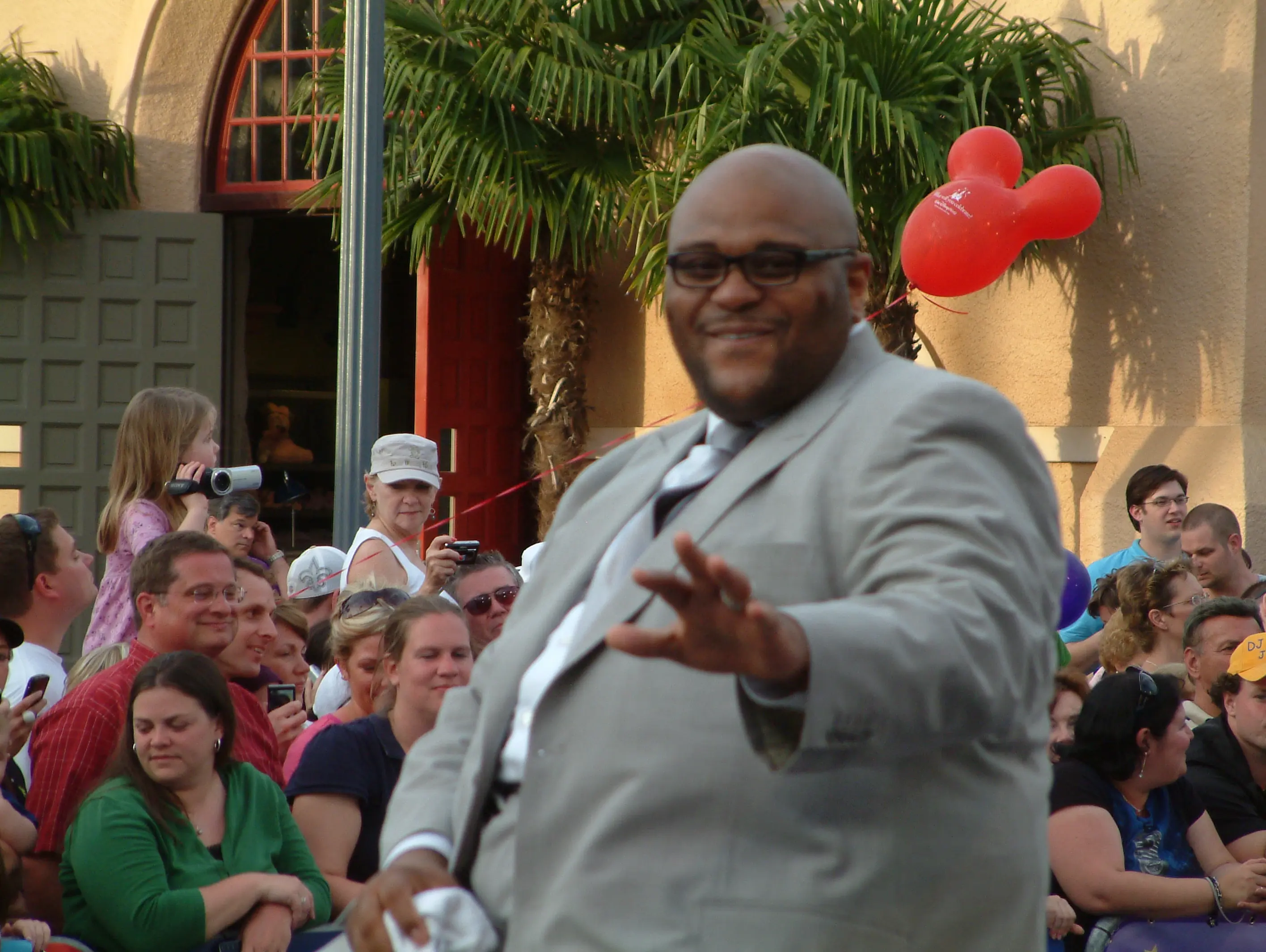 A performer in a grey suit reaching out to a crowd at a theme park, Mickey-shaped balloons behind him