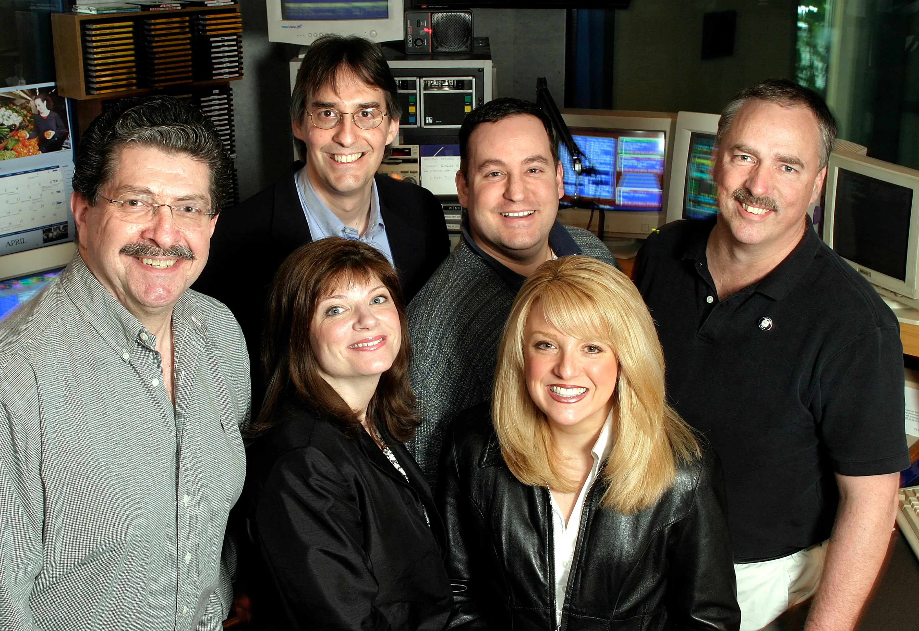 Five people posed together in a broadcast control room, monitors and racks behind them