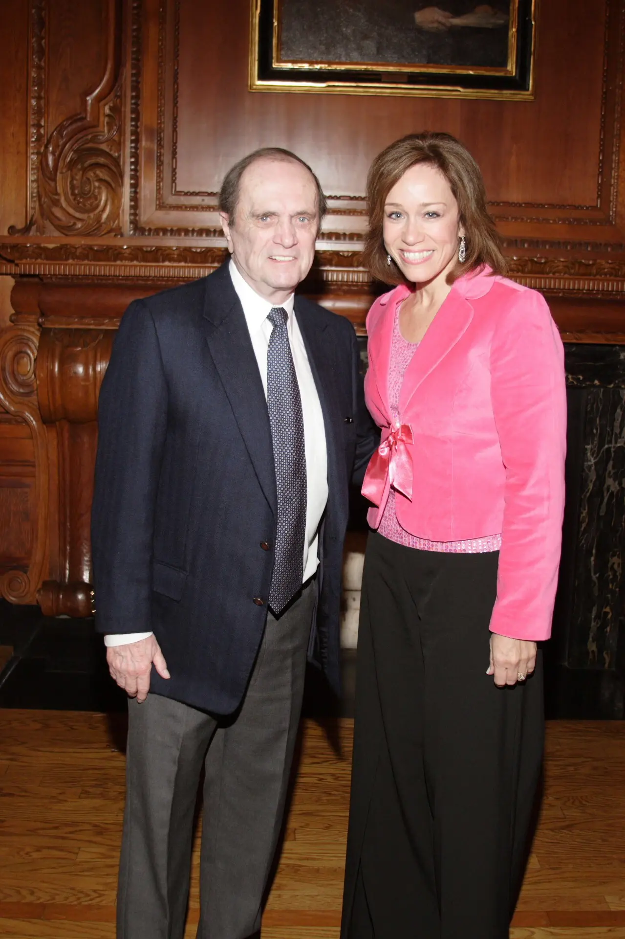 A man in a dark suit and a woman in a pink jacket standing together beside an ornately carved fireplace