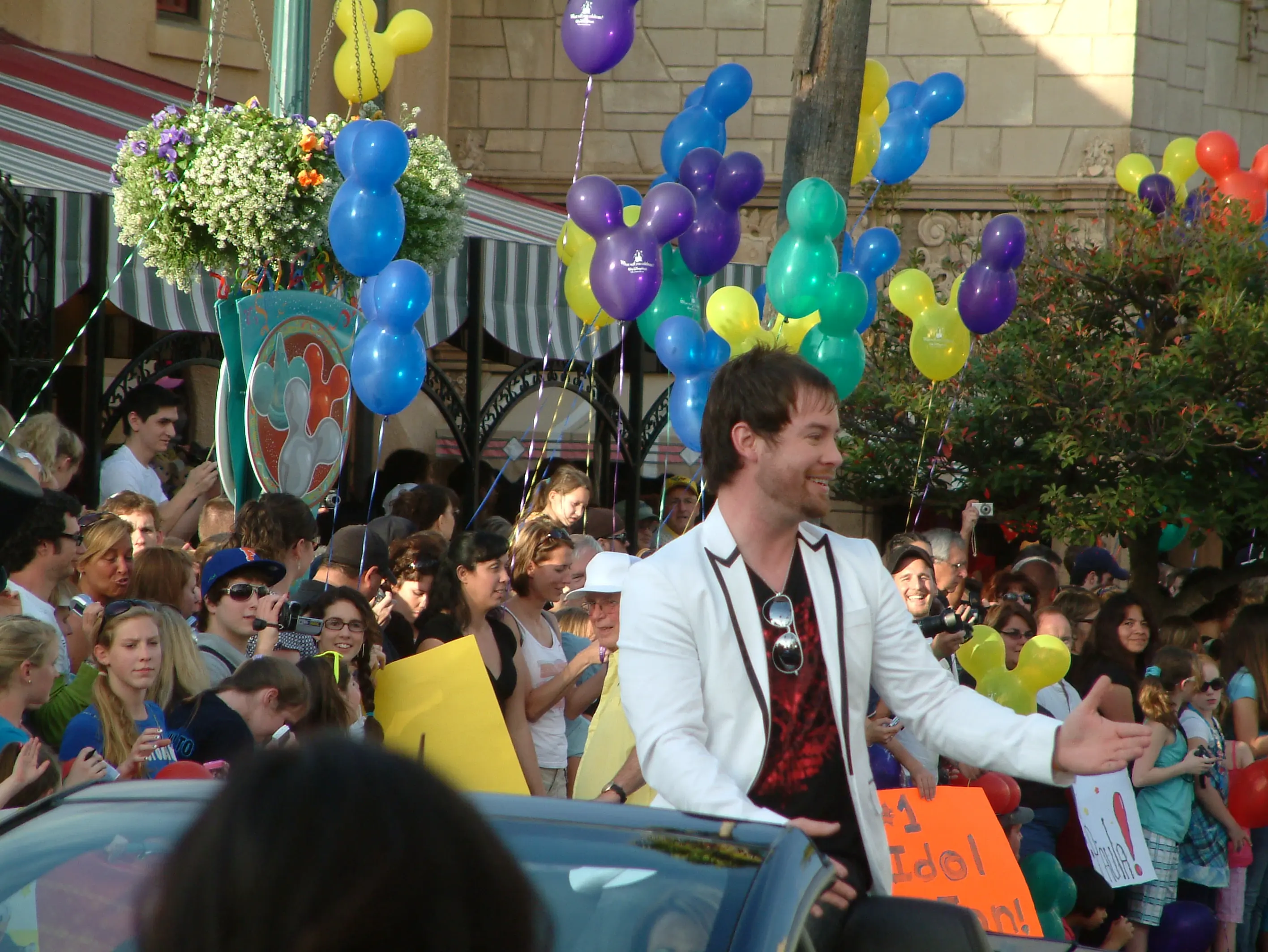 A performer in a white jacket reaching out to fans holding hand-made signs, balloons overhead
