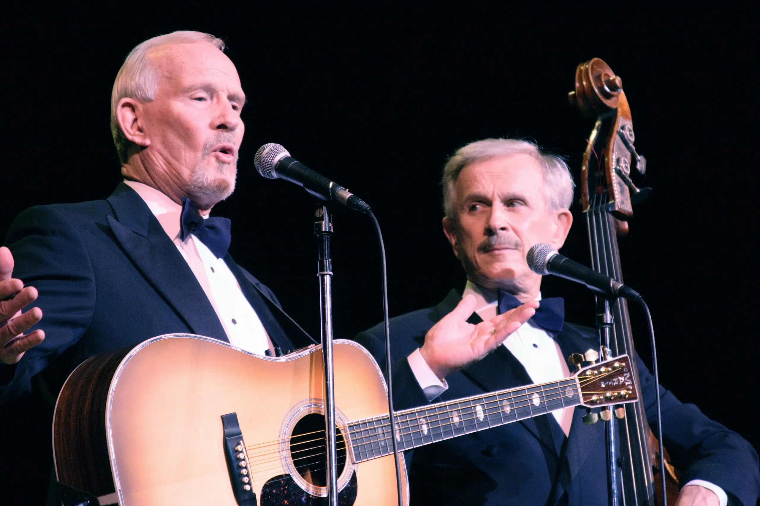 A performer in a tuxedo playing an acoustic guitar at the microphone, a second man in black tie beside him