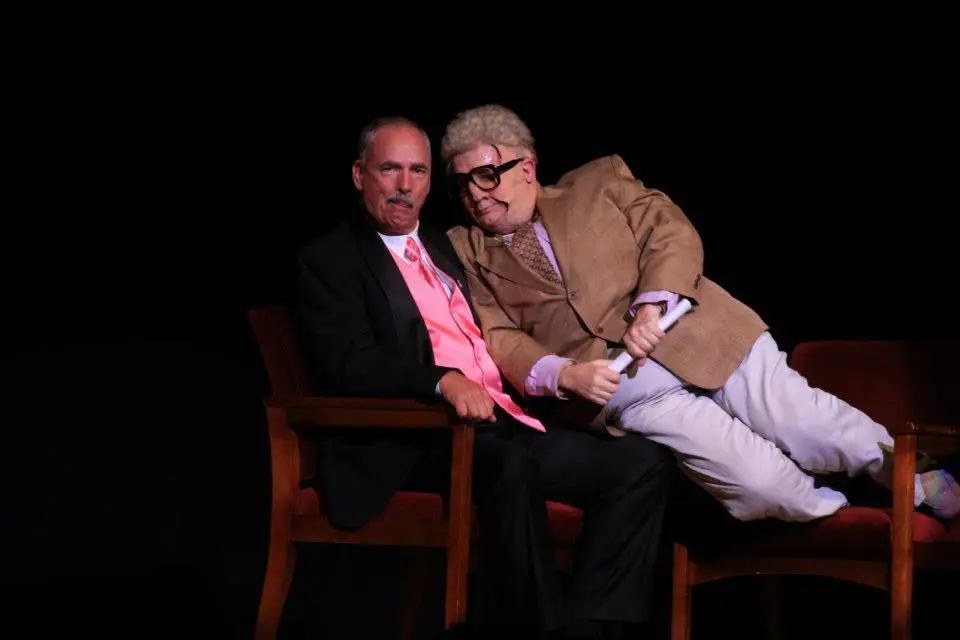 Two performers in a comic bit on stage, one sitting across the other's lap