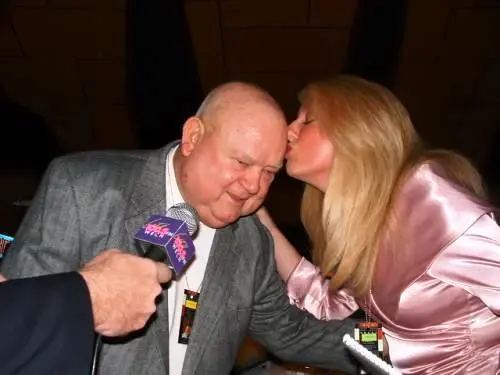 A woman kissing an older man on the cheek as he holds a Lite 100.5 microphone, both laughing