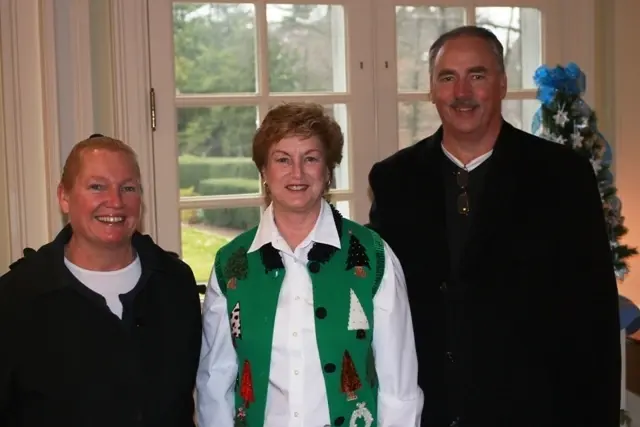 Three people at a holiday gathering, the woman in the centre wearing a green Christmas-tree vest
