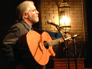 A bearded musician playing acoustic guitar at the microphone beside a lit lantern and brick wall