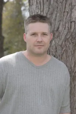 Outdoor portrait of a man in a grey shirt standing against the trunk of a large tree