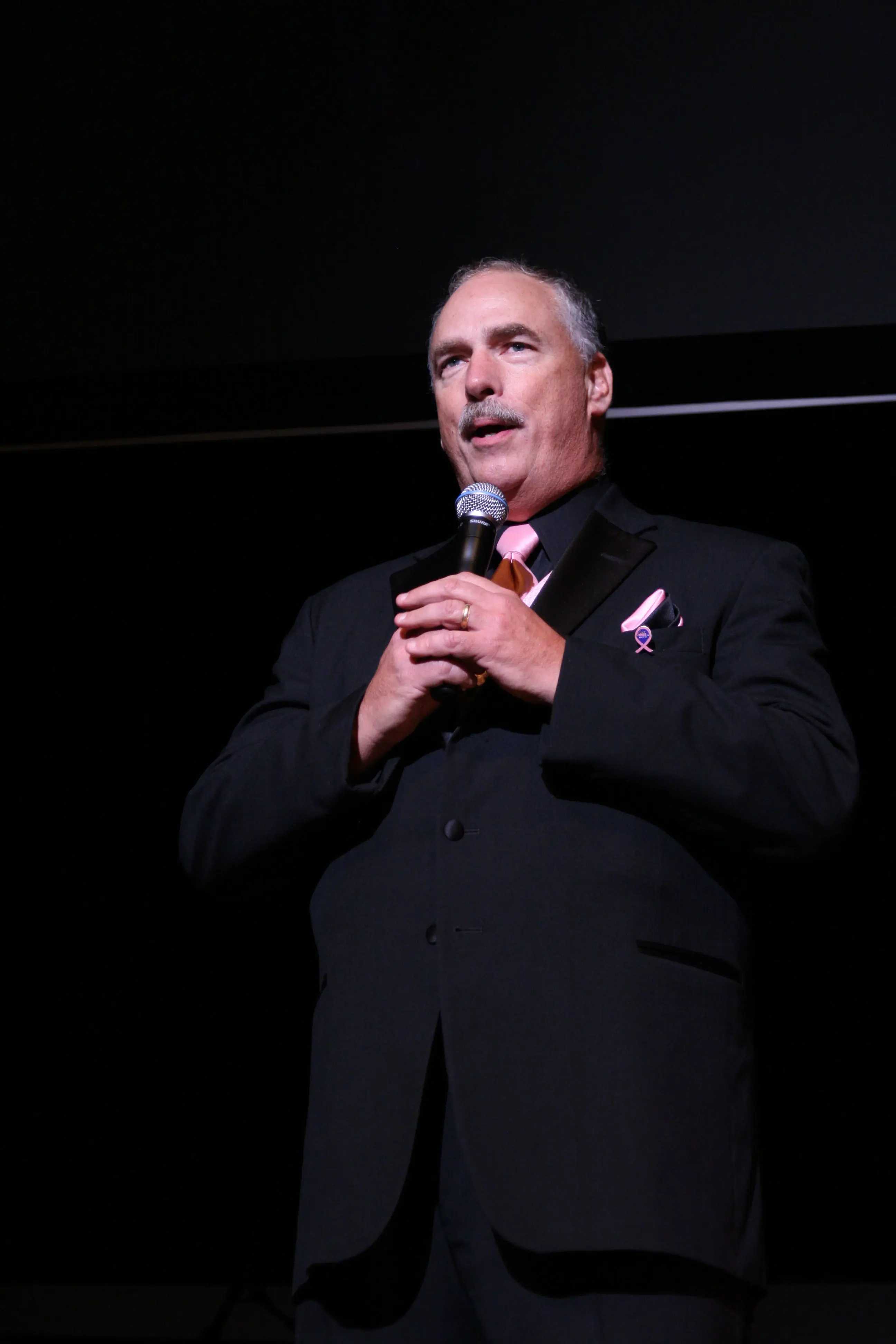 A host in black tie wearing a pink awareness ribbon, speaking into a handheld microphone on a dark stage