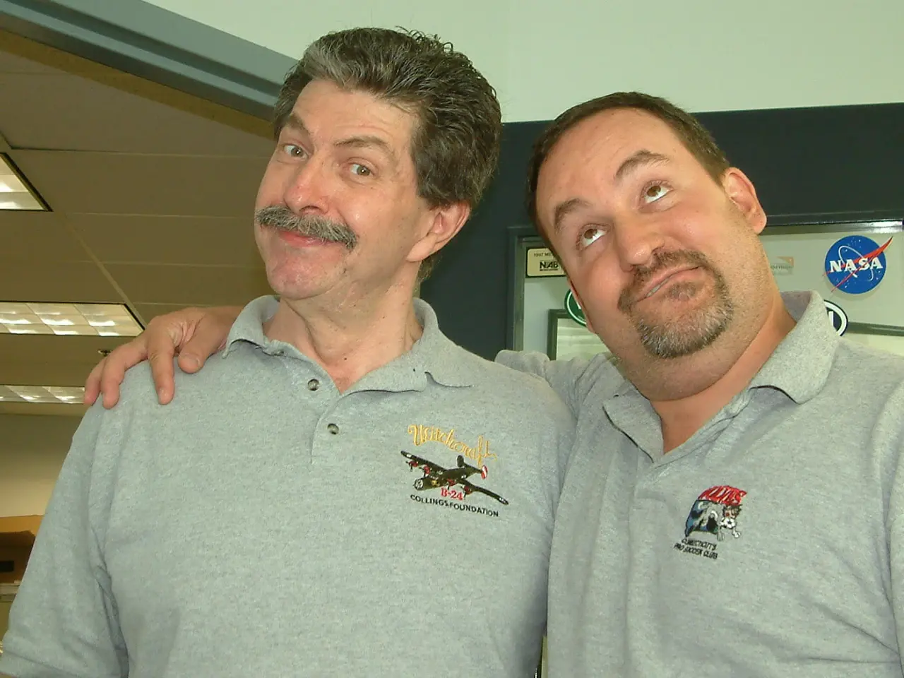 Two men in matching embroidered polo shirts pulling comic faces for the camera in an office