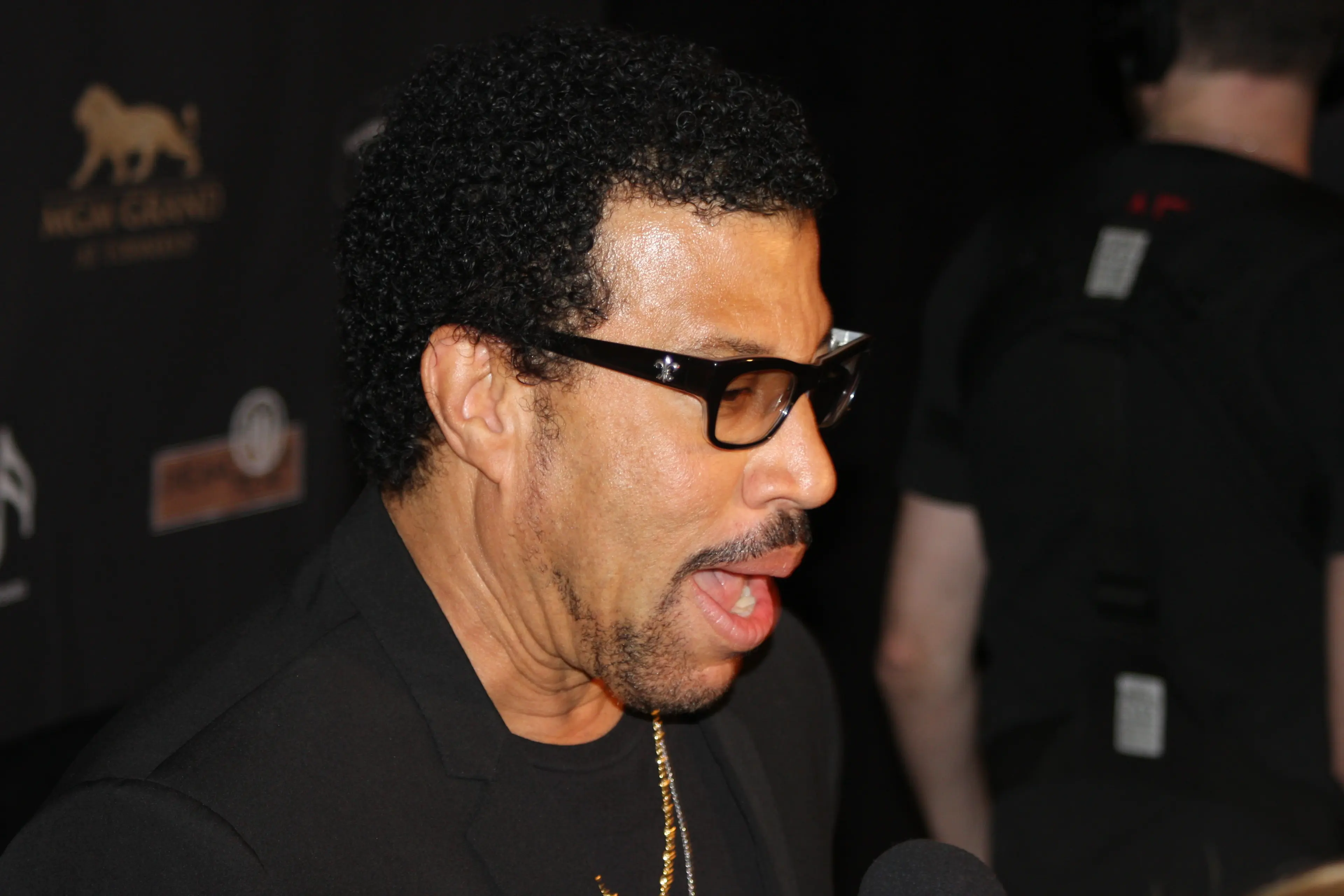 A recording artist in dark-framed glasses speaking at a press event