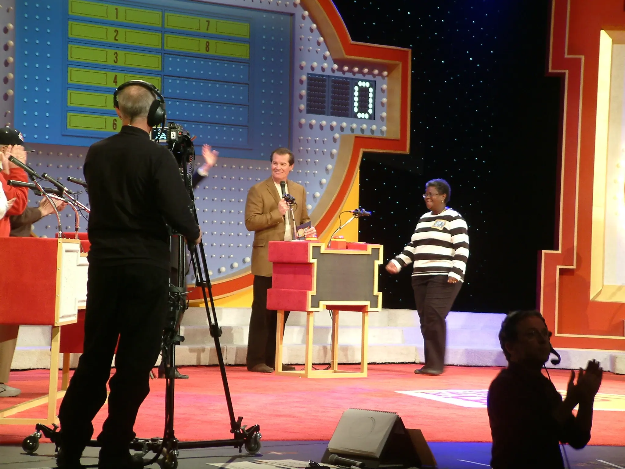 A brightly lit game-show set with a host at the podium and a television camera in the foreground