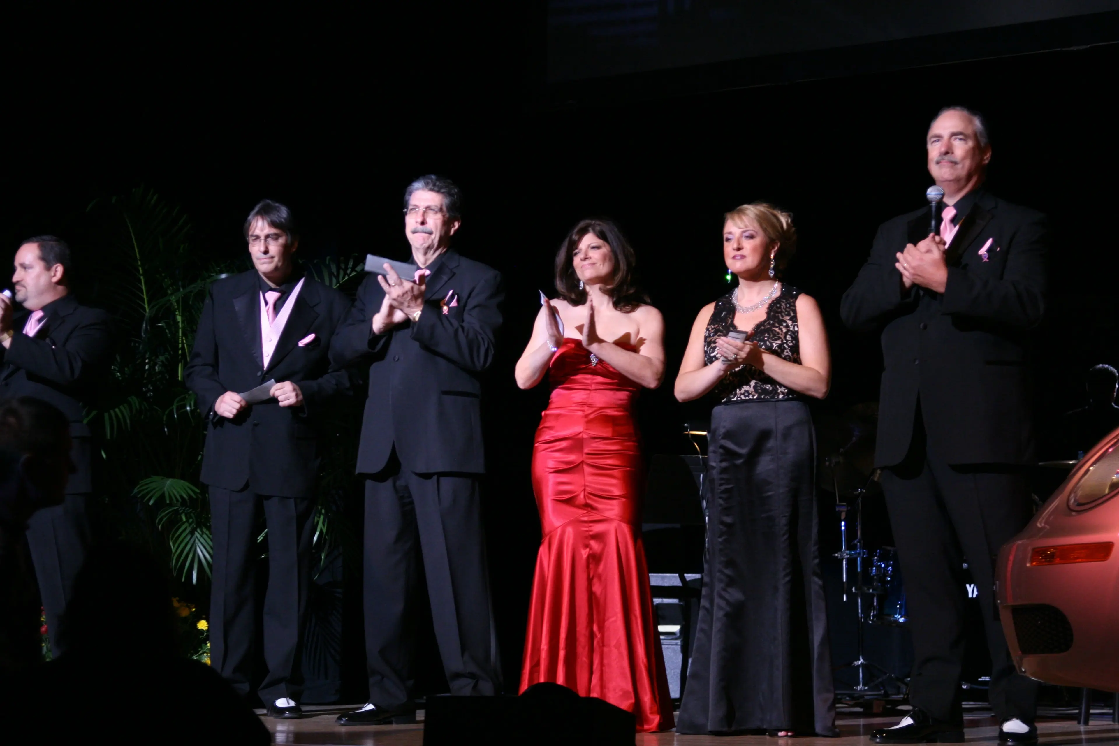 Five people applauding together on stage at a curtain call, one in a long red gown