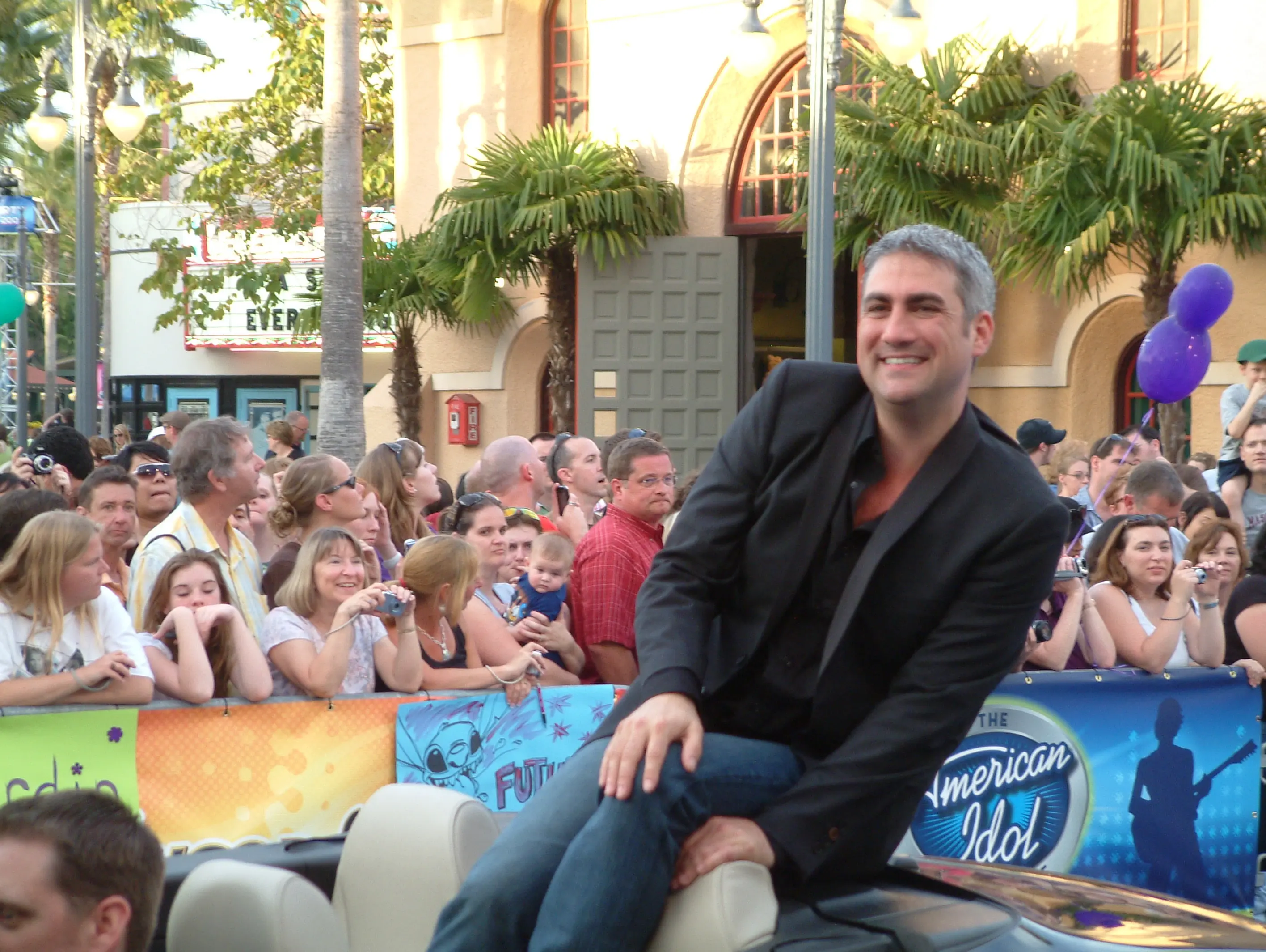 A performer seated in front of an American Idol backdrop with a crowd of onlookers behind the barrier