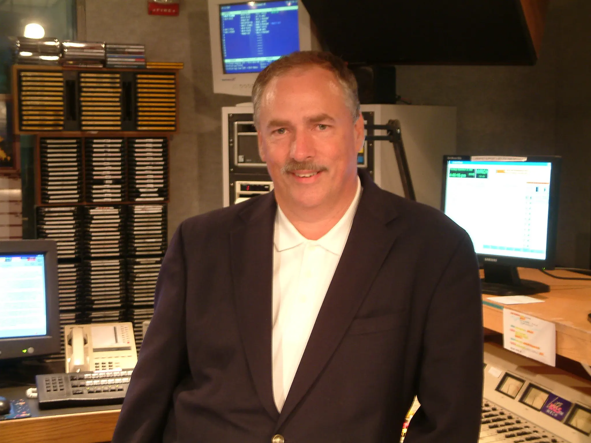 A man in a blazer photographed in the station office, playout monitors on the desk behind him