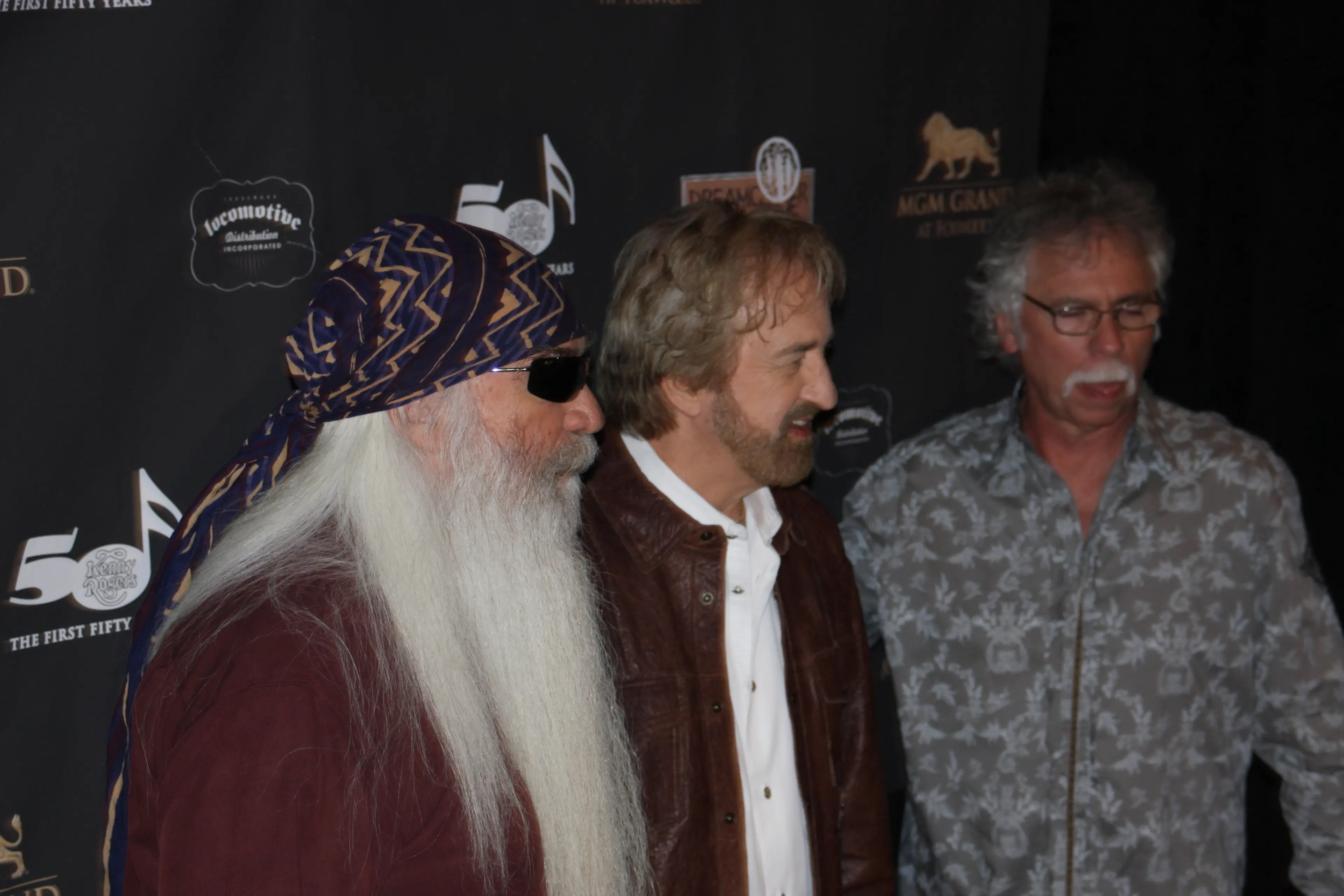 Three band members on a red carpet at an MGM Grand event, one with a long white beard and patterned bandana