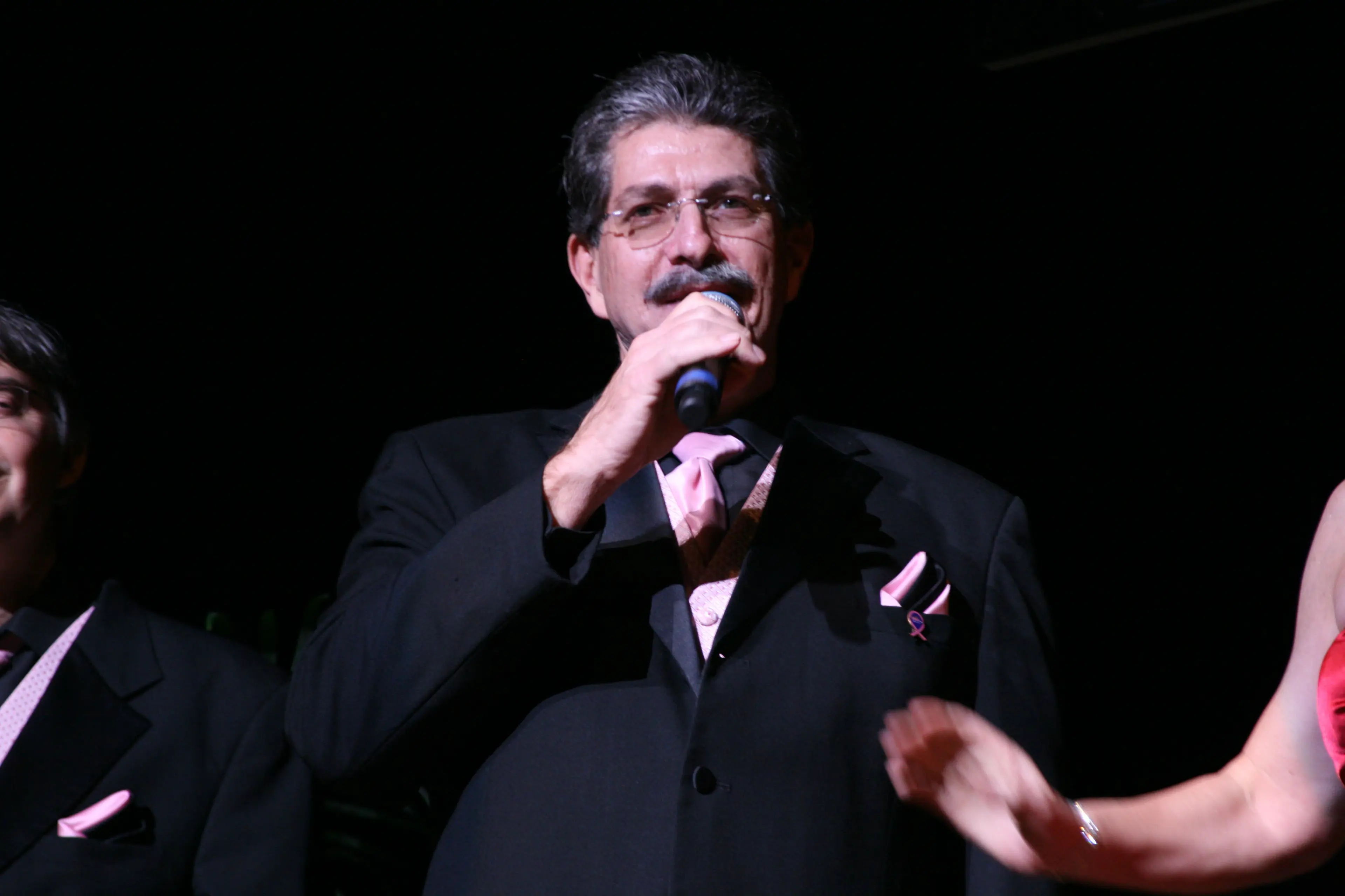 A singer in a tuxedo and pink tie performing into a handheld microphone, hand outstretched to the audience
