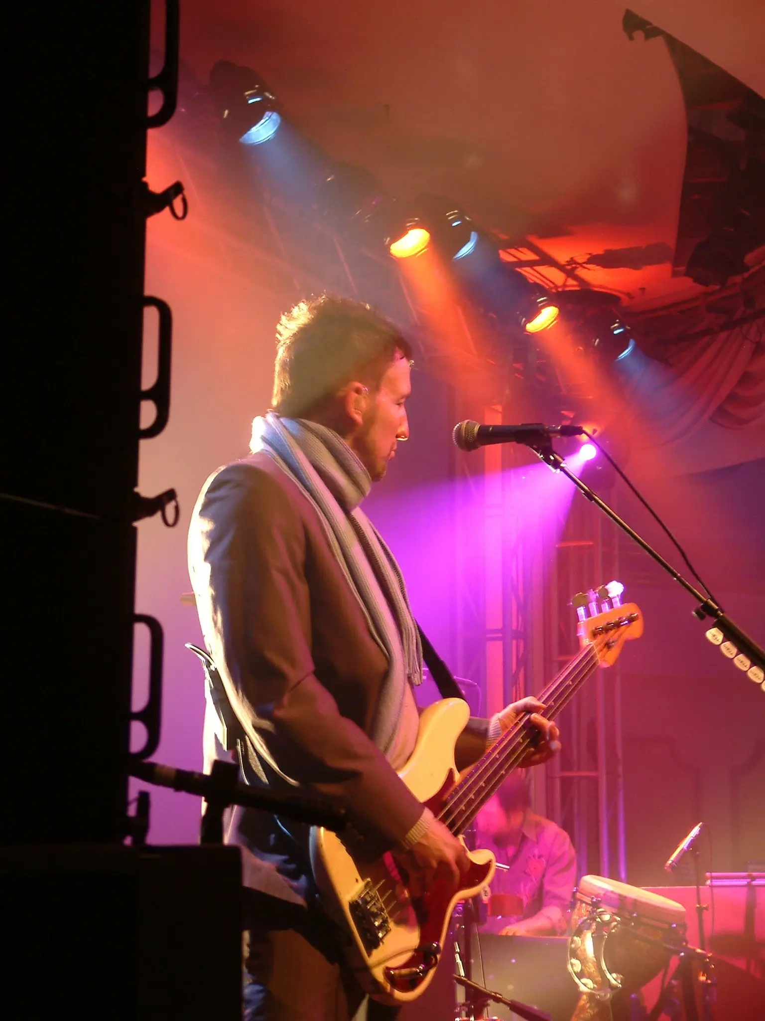 A bass player at the microphone under pink and orange stage lights, scarf around his neck