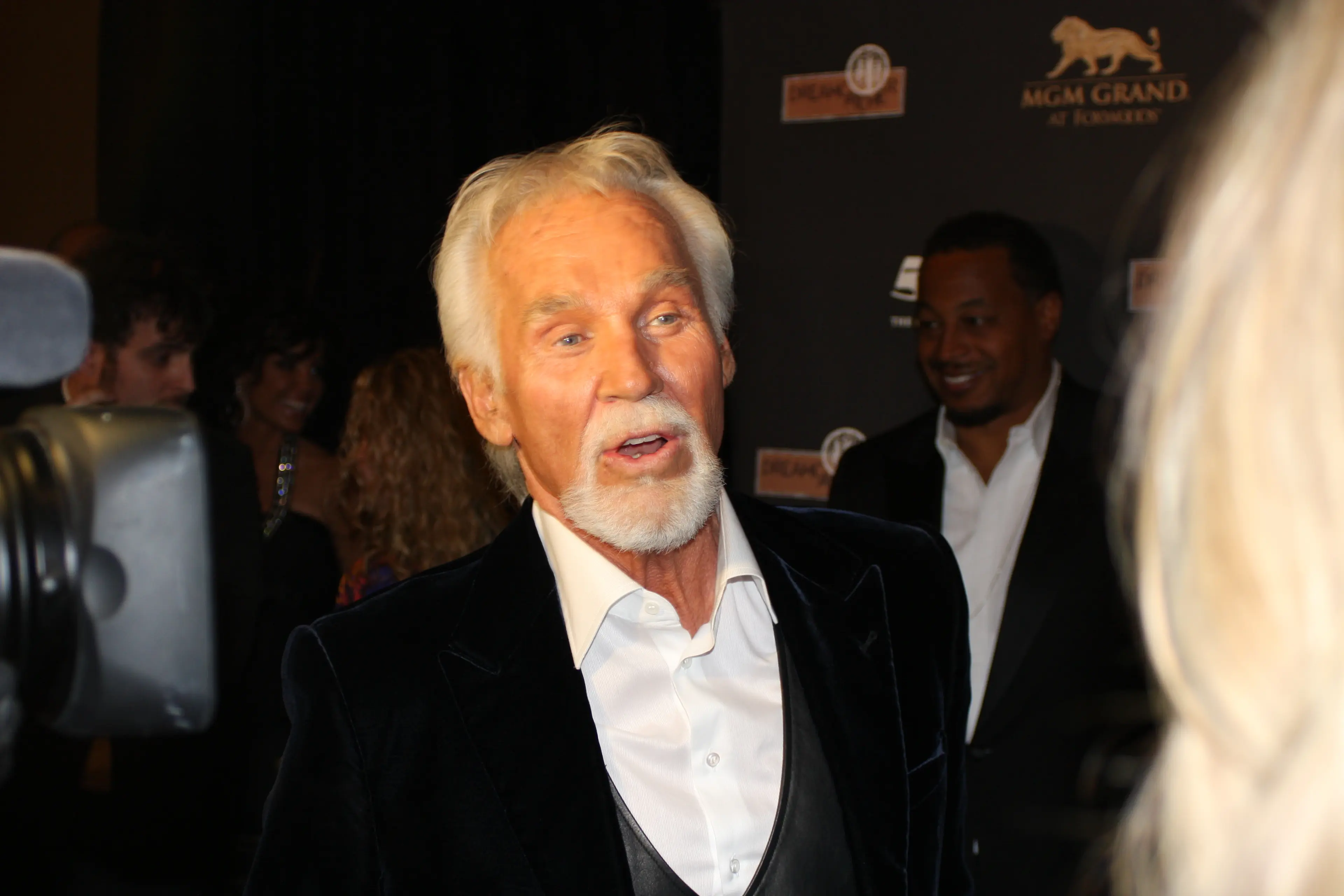 A white-bearded veteran singer in a velvet jacket greeting people on a red carpet at the MGM Grand