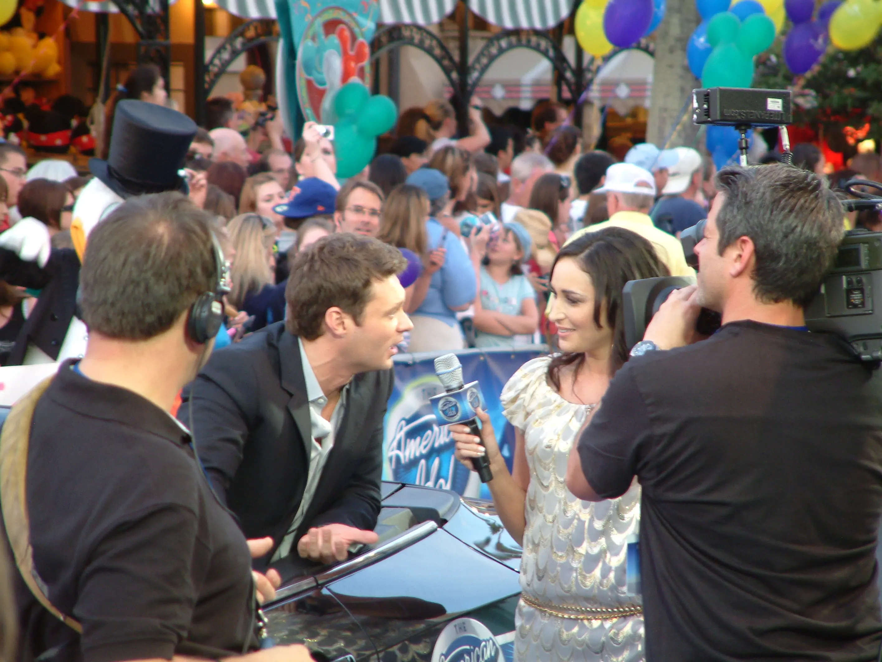 Presenters broadcasting live from an American Idol event with a large crowd pressed behind them