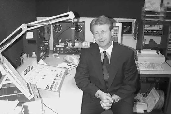 Black and white photograph of a man in a suit beside a vintage radio studio console with reel-to-reel tape decks and a boom microphone