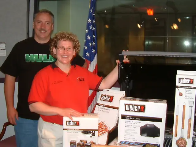 Two people beside a barbecue-grill prize display at a promotional appearance, an American flag behind them