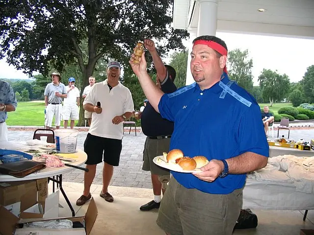 A man in a headband holding up a plate of sliders at an outdoor cookout during a golf outing