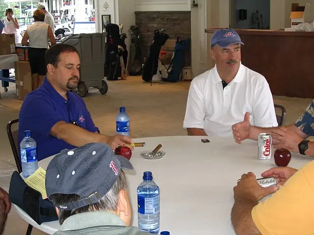 Men seated around a clubhouse table at a golf outing, bags and clubs lined up behind them