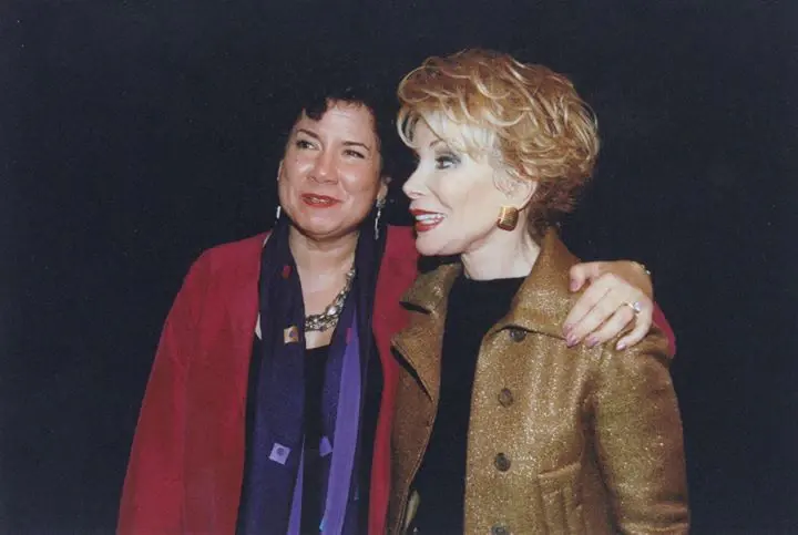 Two women embracing and laughing at an event, one in a red coat and one in gold