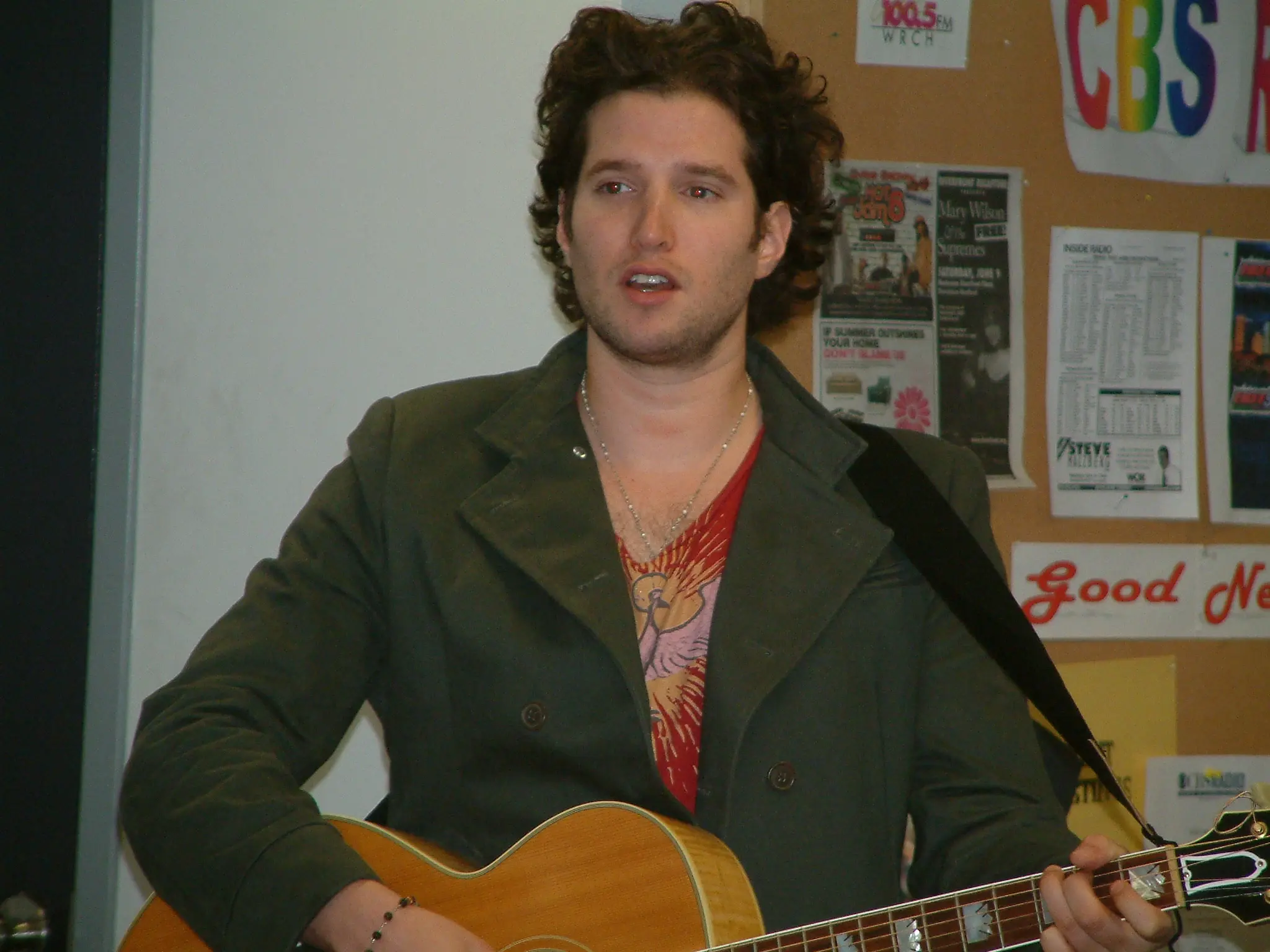 A curly-haired songwriter with an acoustic guitar performing in the station lobby, noticeboard behind him
