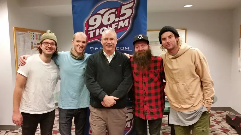 Allan Camp with the members of a visiting band in the studio, a 96.5 TIC-FM banner on the wall behind them